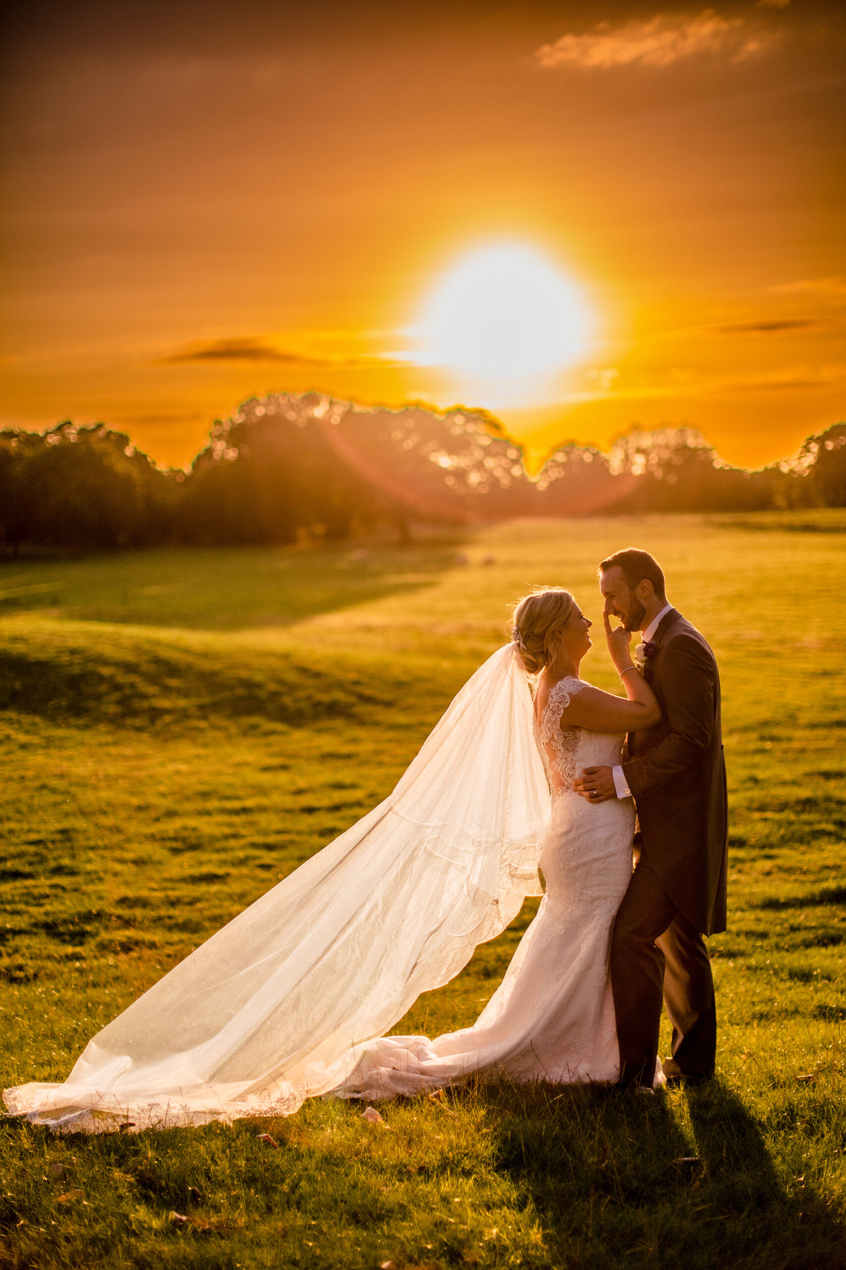 Bride and groom embracing in the golden sunset light with her long veil across the grass, Cheshire wedding photographer