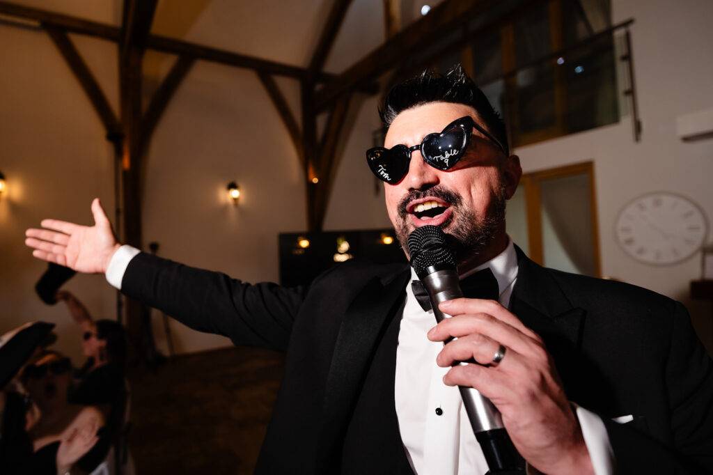 A man in heart shaped sunglasses on the microphone with his arm out, geeing up the room, Nottingham wedding photographer