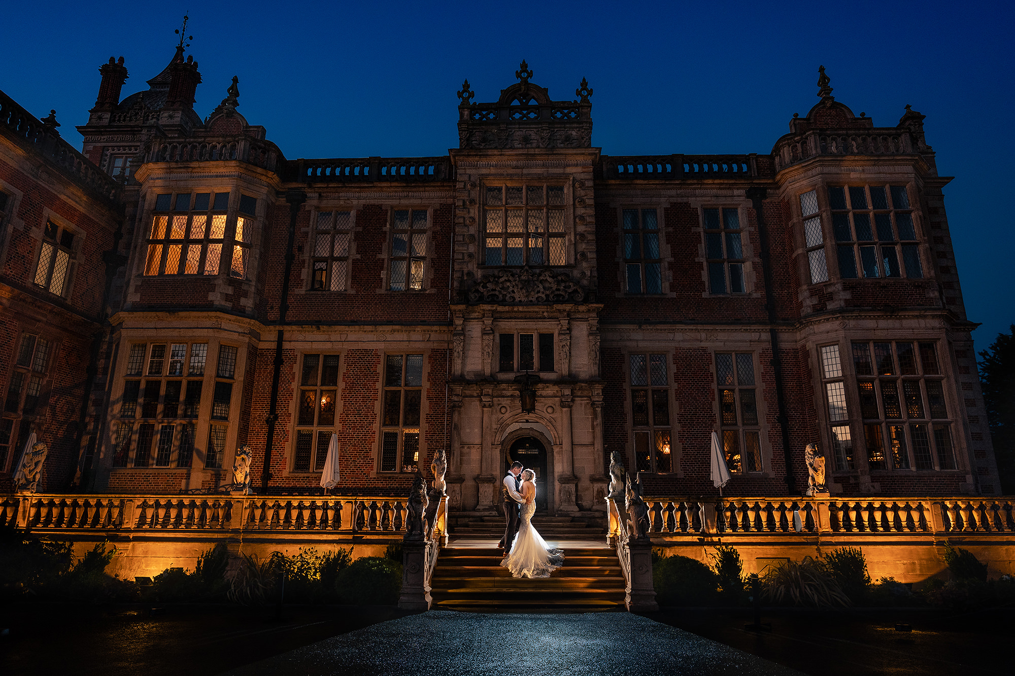 Crewe Hall lit up against a deep blue evening sky with the bride and groom tiny in the lit doorway at the top of the steps