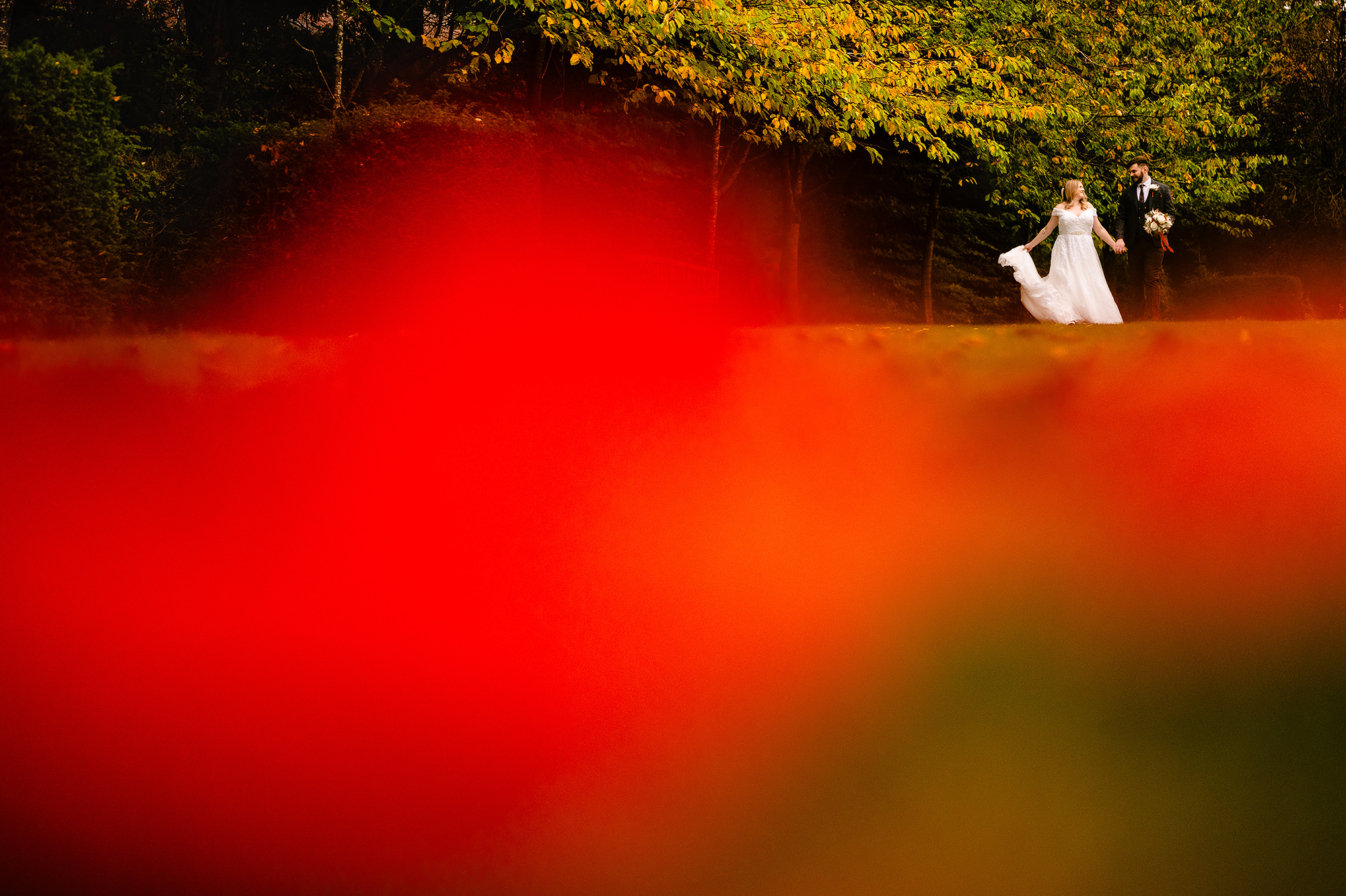 Annie and Tom walking hand in hand through the grounds, shot through a foreground of autumn leaves, Crewe Hall wedding photographer