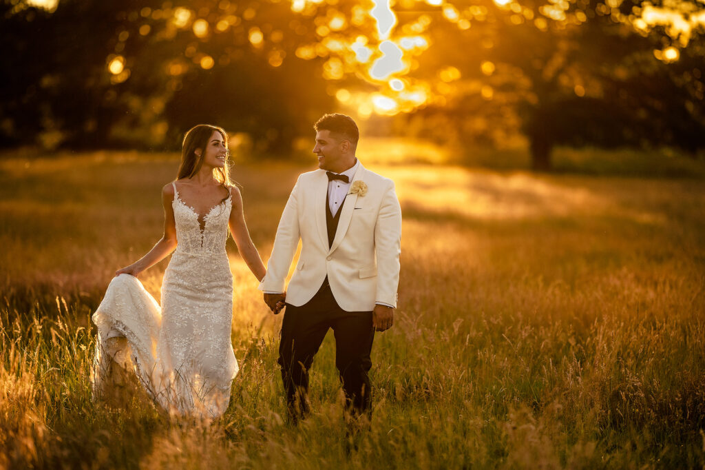 Gemma and Tyrone walking hand in hand through golden long grass at sunset, Merrydale Manor wedding photographer