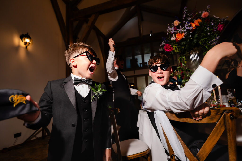 Two boys in black tie and sunglasses cheering and waving napkins, Swancar Farm wedding photographer