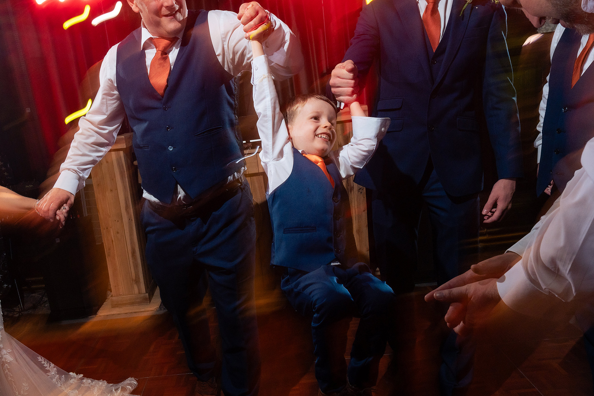 A small boy in a navy suit being swung off his feet between two men on the dance floor under coloured lights