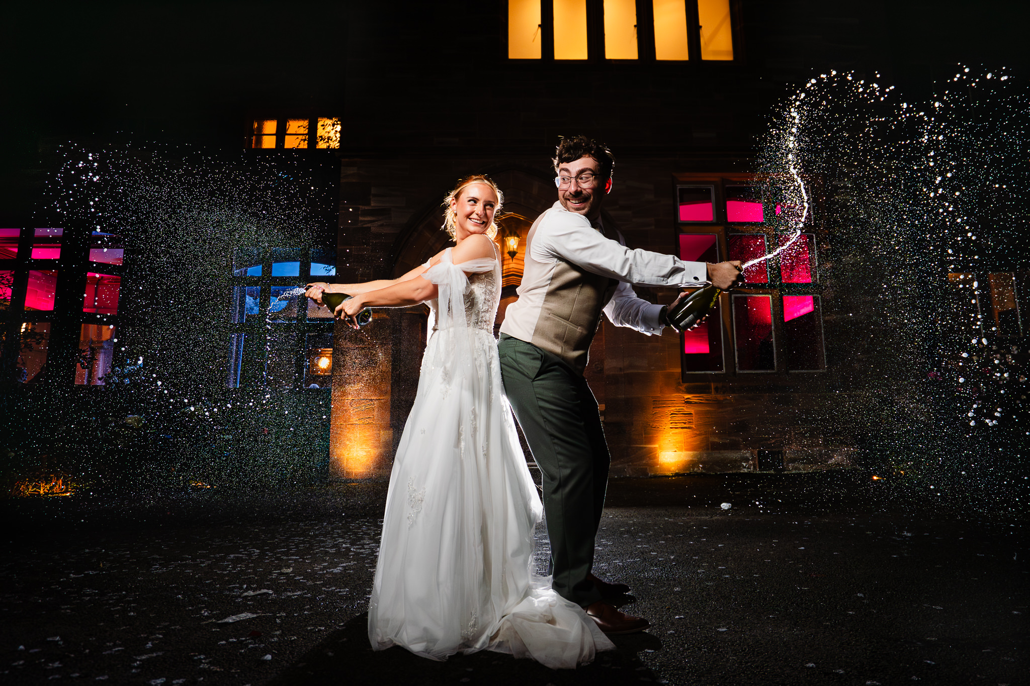 Bride and groom spraying champagne at night in front of the lit up Salt House