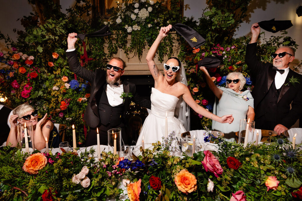 The top table waving napkins in sunglasses in front of a huge floral installation, Swancar Farm wedding photographer