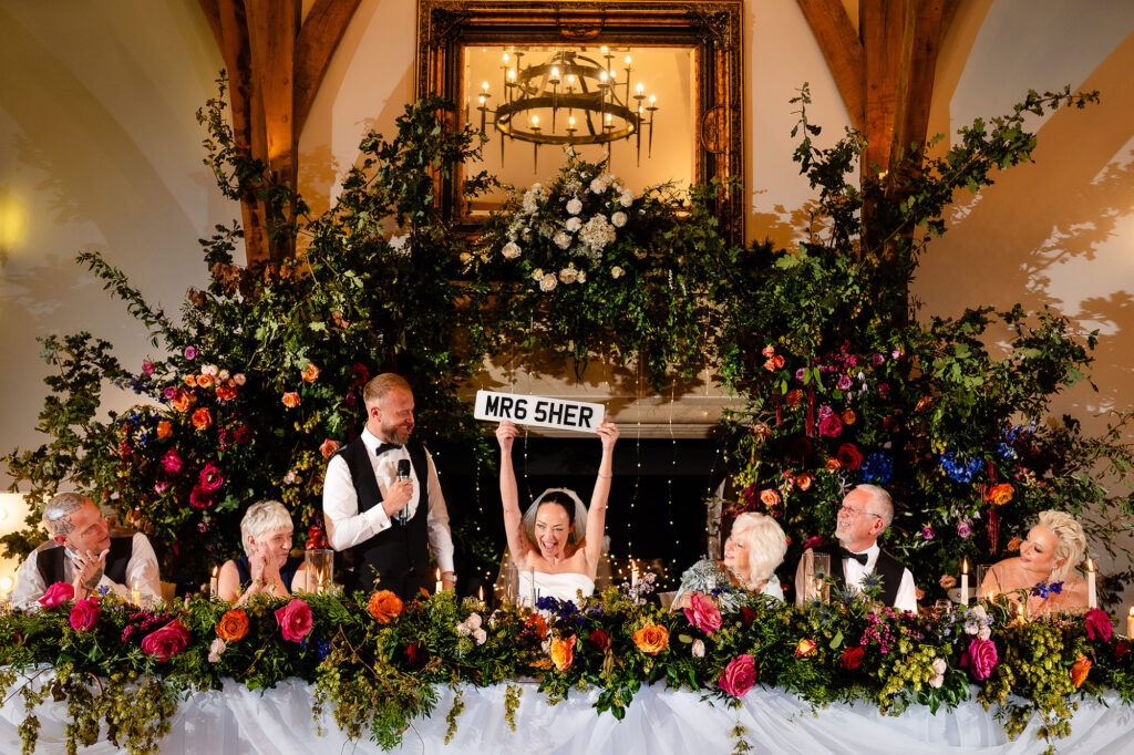 Kim holding a MR6 SHER number plate over her head while Glynn speaks at the top table, Swancar Farm wedding photographer