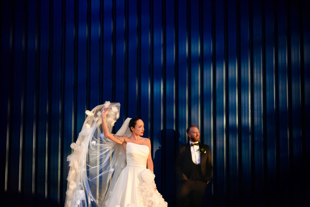 Kim flicking her rose covered veil against a deep blue wall in late sun as Glynn watches, Swancar Farm wedding photographer
