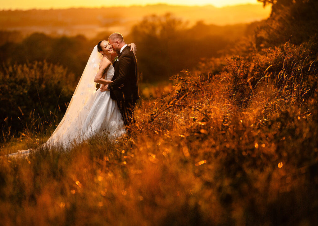 Kim and Glynn embracing in long golden grass at sunset, Swancar Farm wedding photographer