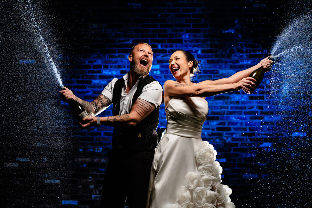 Kim and Glynn spraying champagne against a blue lit brick wall at the end of the night, Swancar Farm wedding photographer