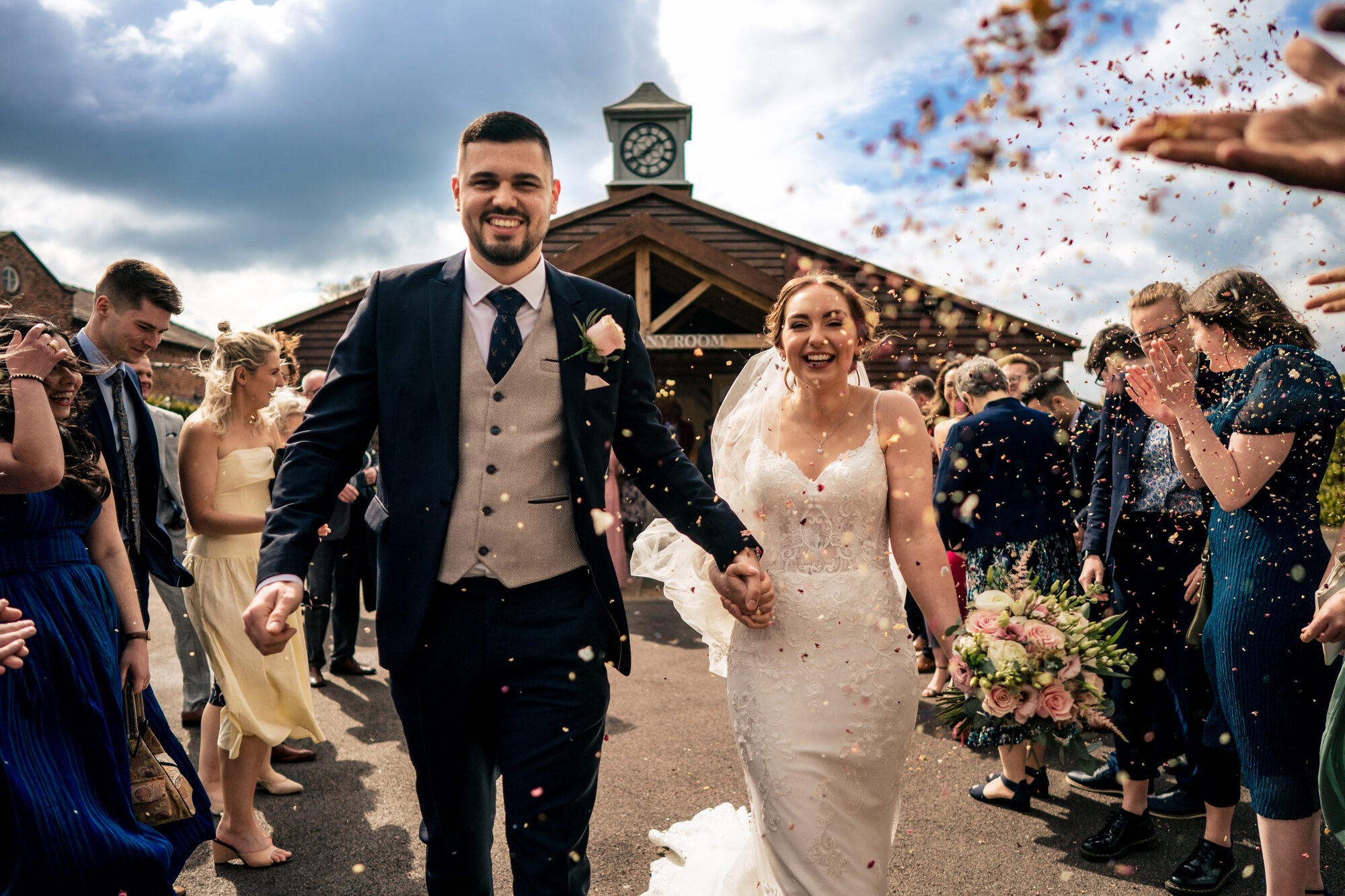 Newlyweds walking through confetti outside a barn with a clock tower