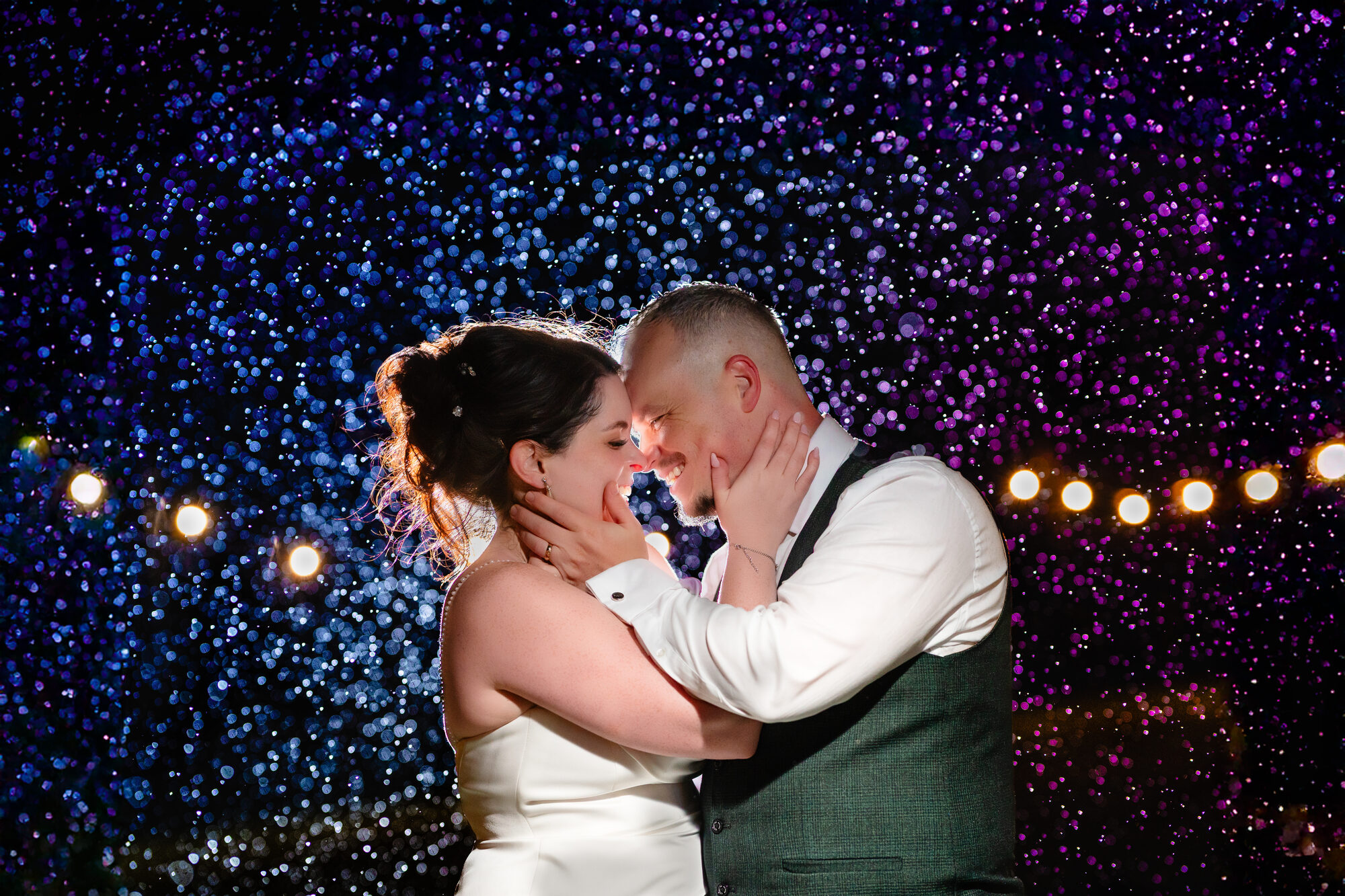 Bride and groom close together against a wall of blue fairy lights