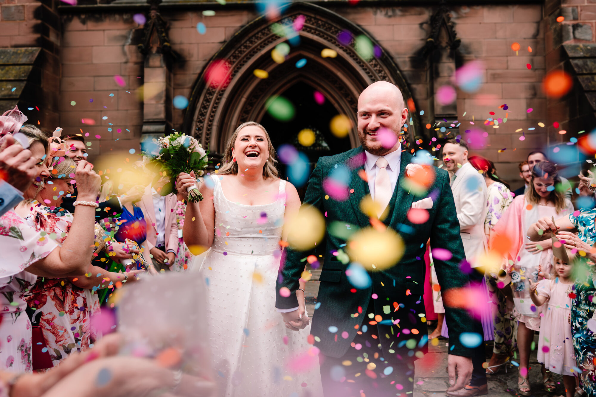 Bride and groom laughing in a shower of brightly coloured confetti outside church