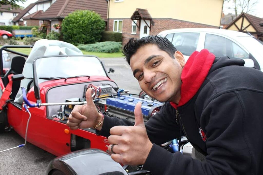 Dev grinning and giving two thumbs up next to a red open top sports car
