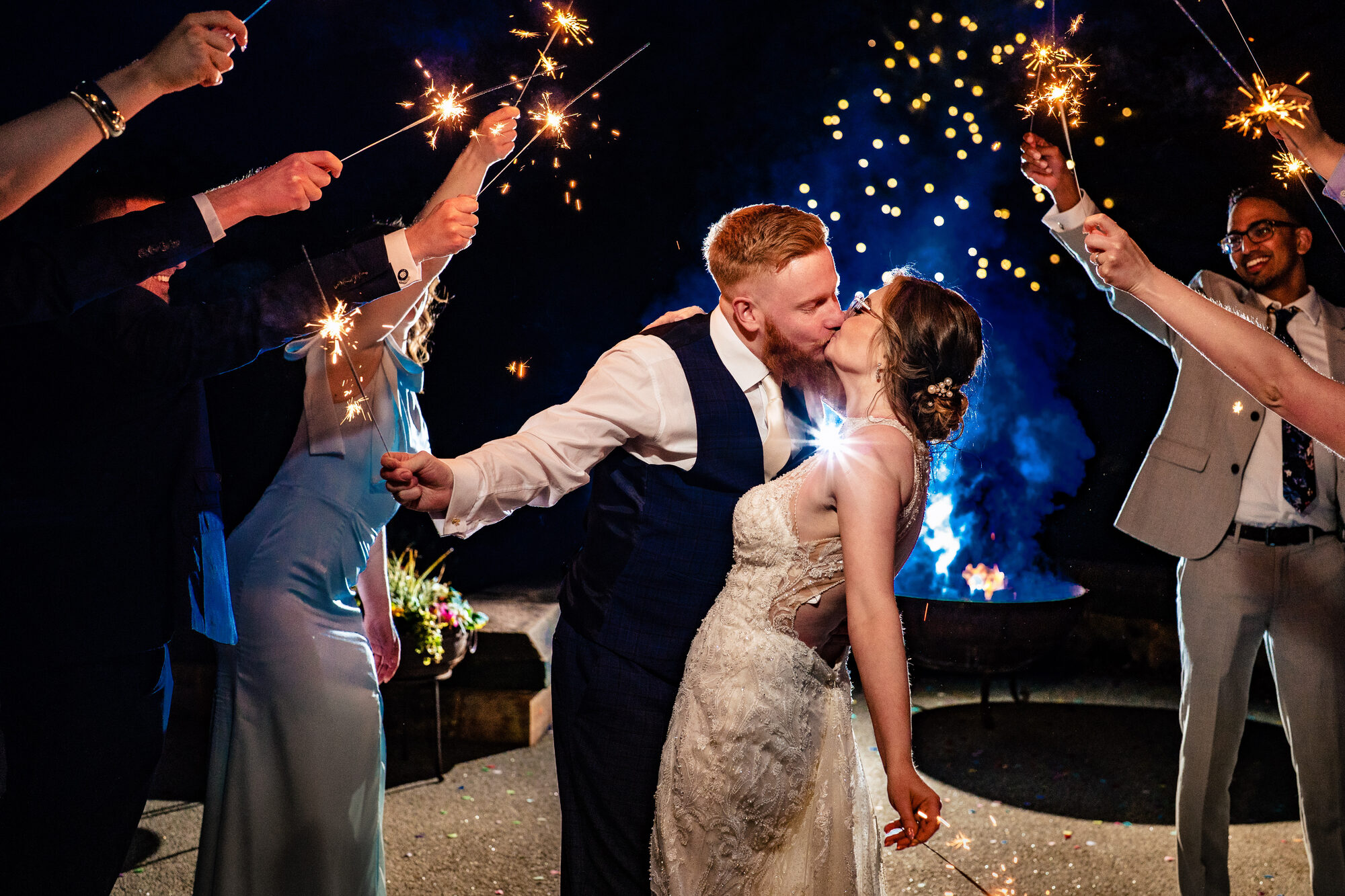 Bride and groom kissing surrounded by sparklers beside a fire pit