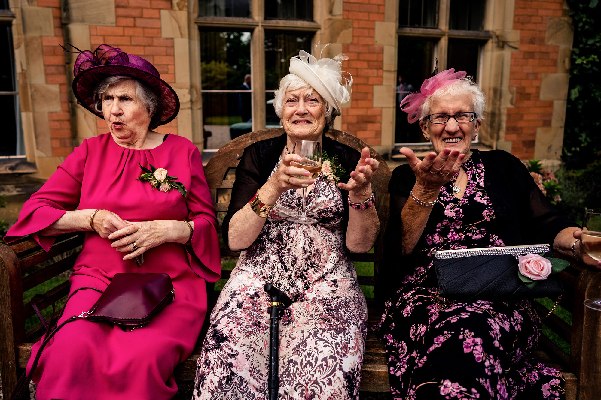Three older wedding guests laughing together on a bench with their drinks