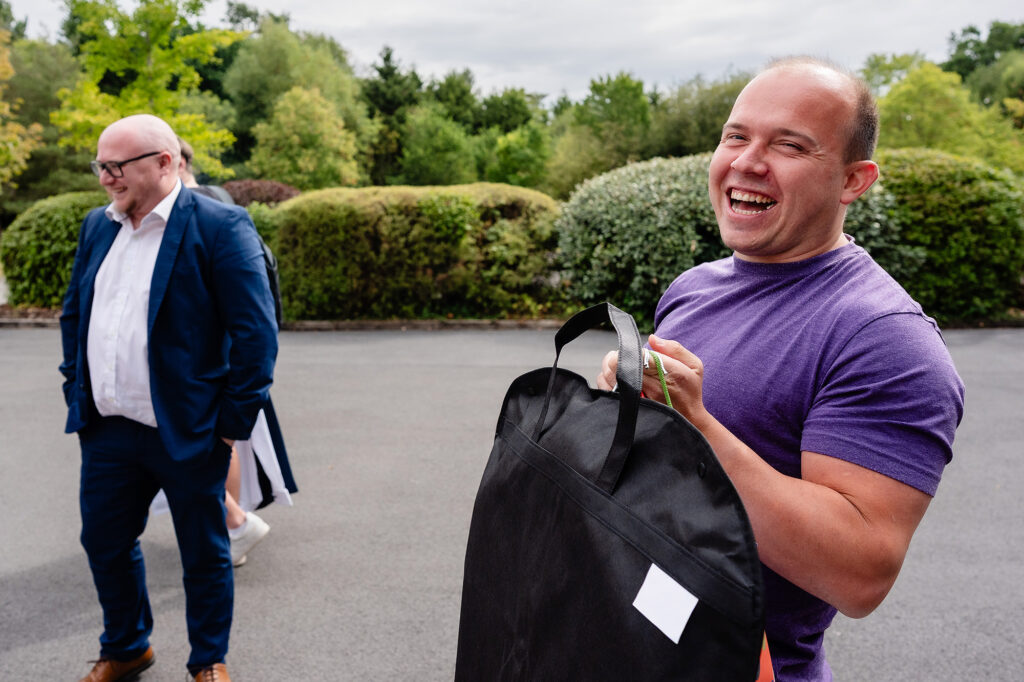 Groom in a purple t-shirt laughing as he carries his suit bag outside The Oak Tree of Peover on the wedding morning