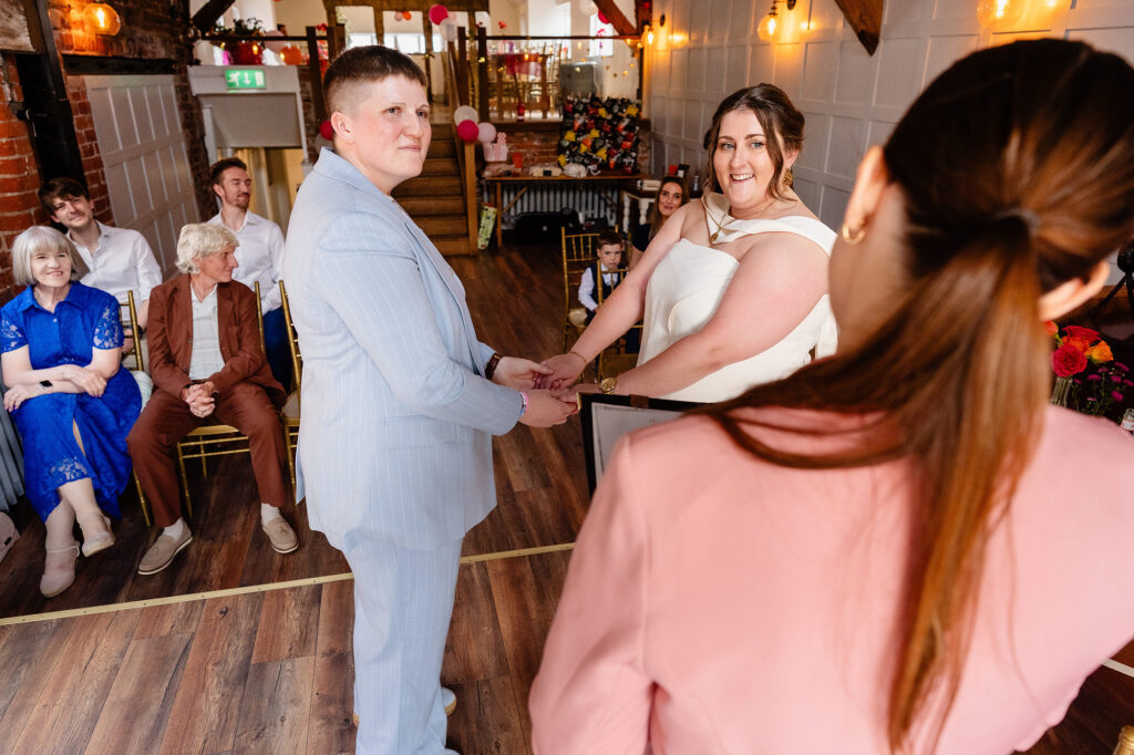 Marianne in a pale blue suit and Emma in a cream jumpsuit holding hands at the start of their ceremony at The Hidden Barn @ 94 in Knutsford