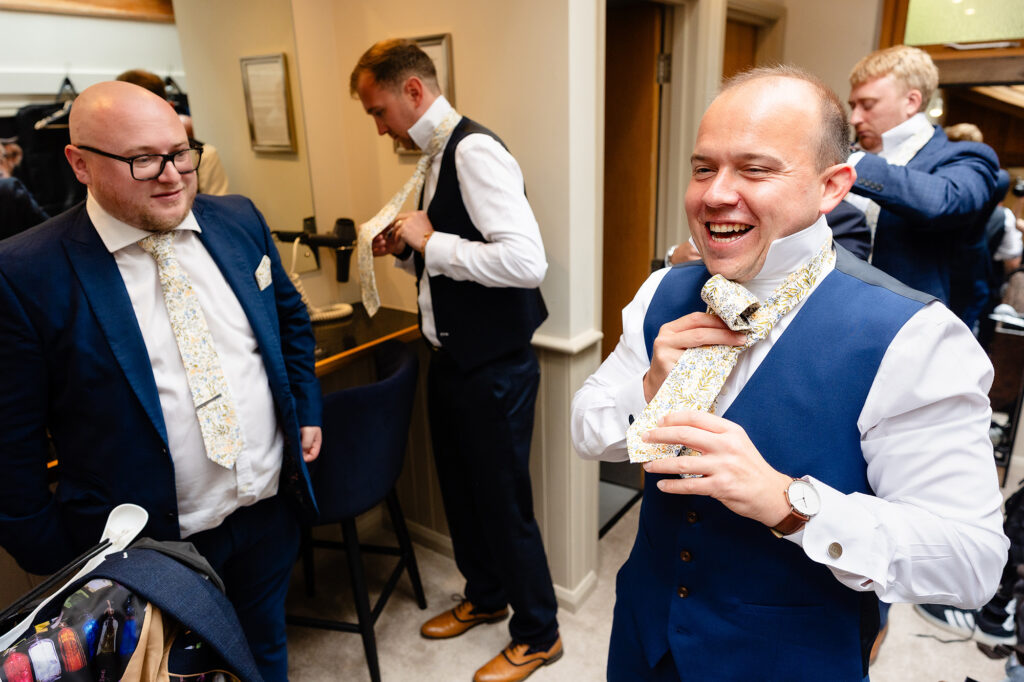 Groom laughing as he ties his floral tie while his groomsmen get dressed around him, Cheshire wedding photographer