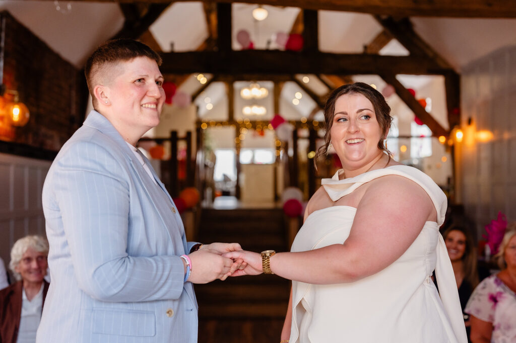 Marianne and Emma holding hands and smiling during their wedding ceremony under exposed wooden beams