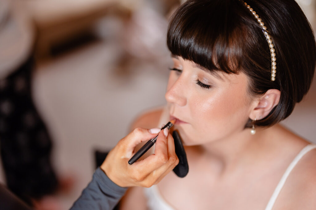 Bride with a fringed bob and pearl headband having her lip liner applied on the wedding morning