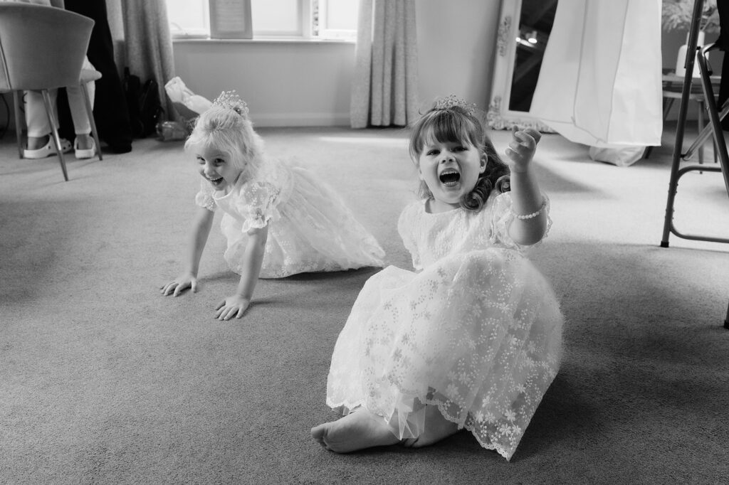 Black and white photo of two flower girls in lace dresses playing on the bedroom floor, one shrieking with laughter