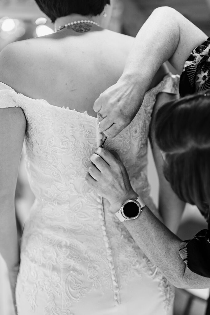 Black and white close up of hands fastening the buttons down the back of a lace wedding dress