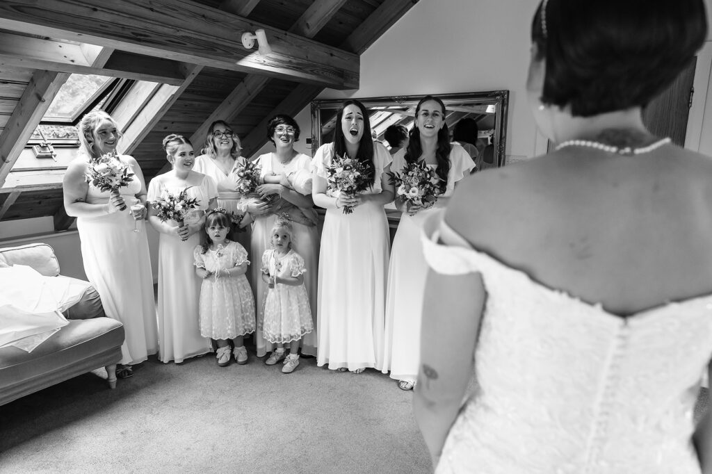Black and white photo of bridesmaids and flower girls gasping as they see the bride in her dress for the first time