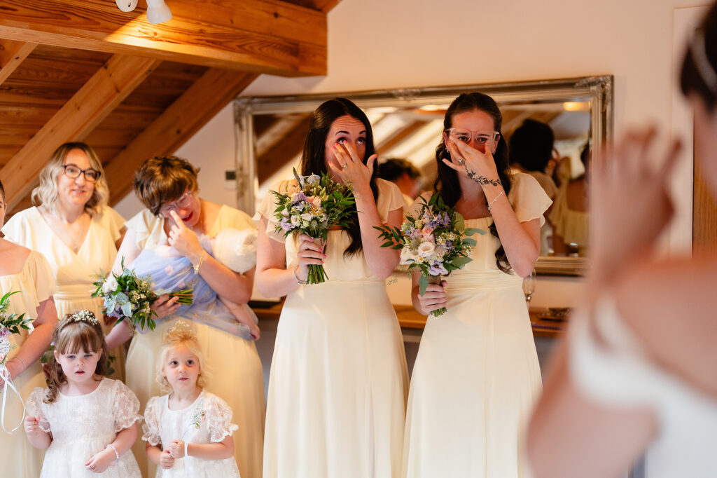 Bridesmaids in pale yellow dresses with hands over their faces, tearful at their first look at the bride