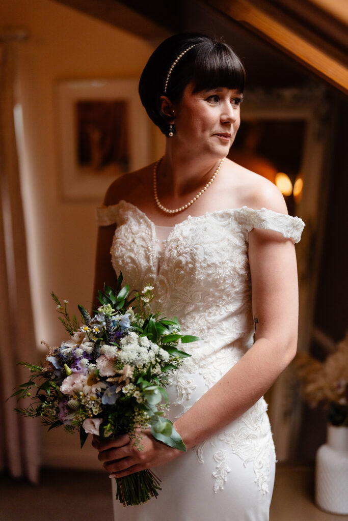 Bride in an off the shoulder lace dress and pearl necklace holding her wildflower bouquet in the loft room, Cheshire wedding photographer