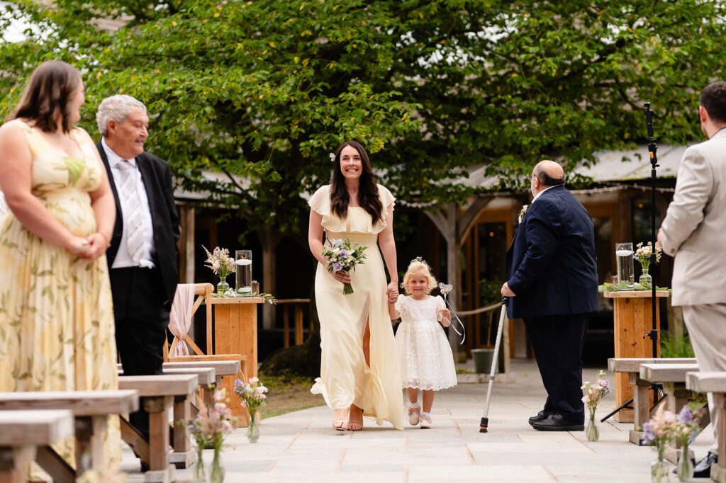 Smiling bridesmaid walking a flower girl down the outdoor ceremony aisle at The Oak Tree of Peover