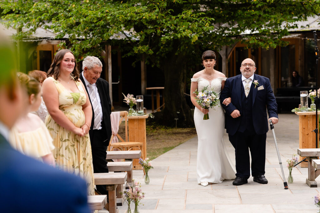 Bride walking down the outdoor aisle arm in arm with a guest in a navy suit, past wildflower jars on the benches