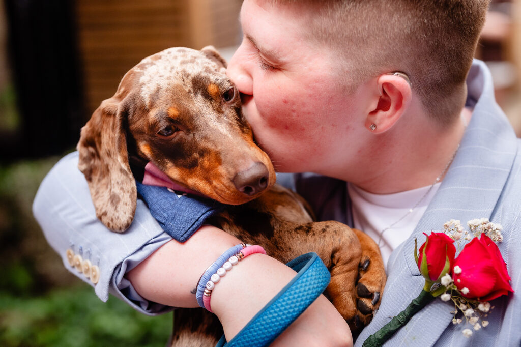 Marianne kissing her dapple sausage dog, a red rose buttonhole on her suit