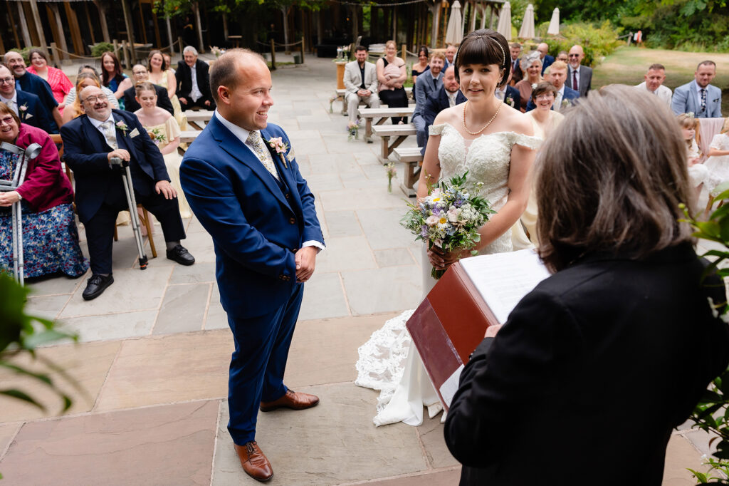 Bride and groom at the front of the outdoor ceremony with guests on wooden benches behind them, Oak Tree of Peover wedding