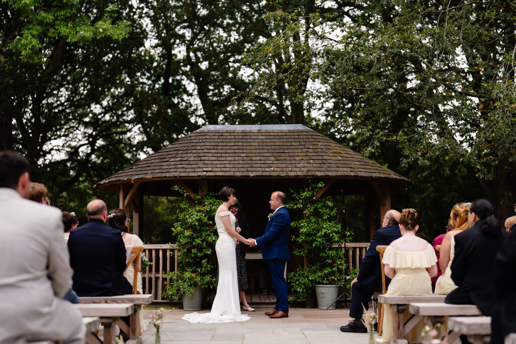 Wide view of the couple holding hands beneath the wooden gazebo in the garden ceremony space at The Oak Tree of Peover