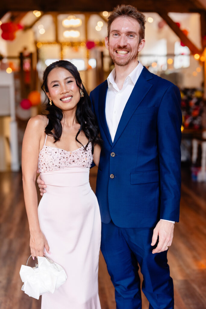 Two wedding guests in a pink dress and a navy suit posing inside The Hidden Barn @ 94