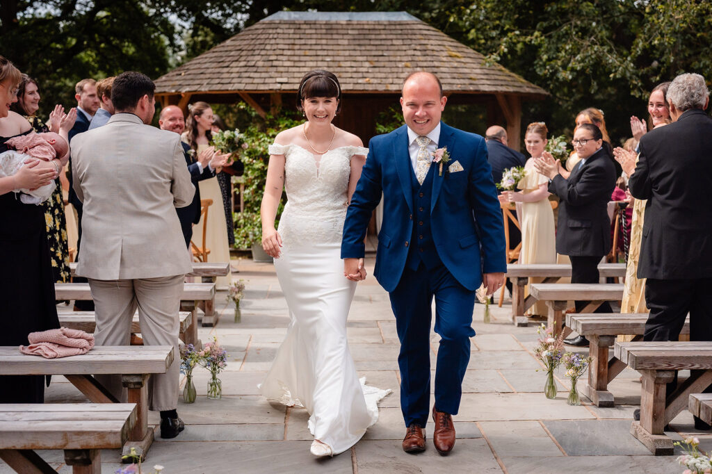Newlyweds grinning as they walk back up the aisle hand in hand while guests clap