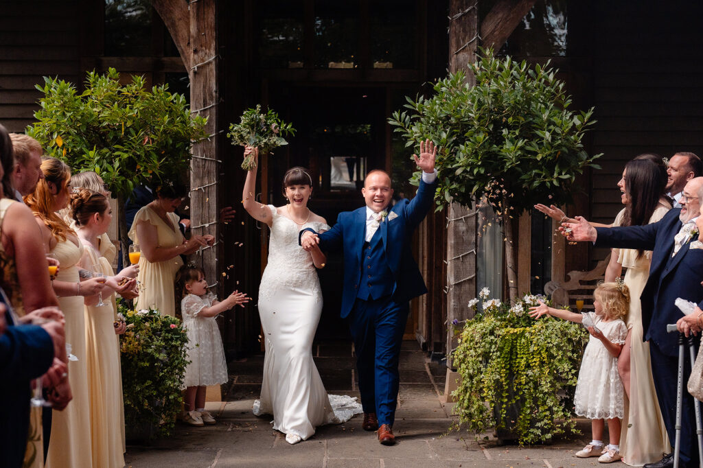 Bride and groom raise their arms as flower girls and guests throw confetti outside the front of The Oak Tree of Peover