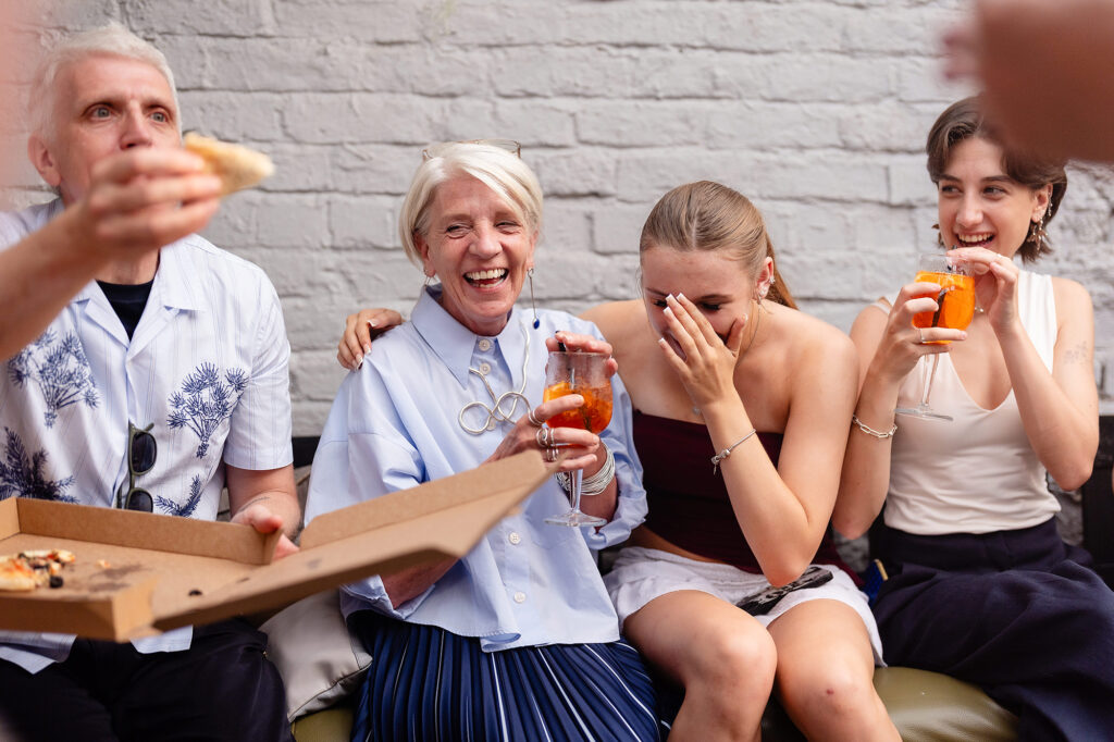 Guests laughing on a sofa with boxes of pizza, one holding up a slice