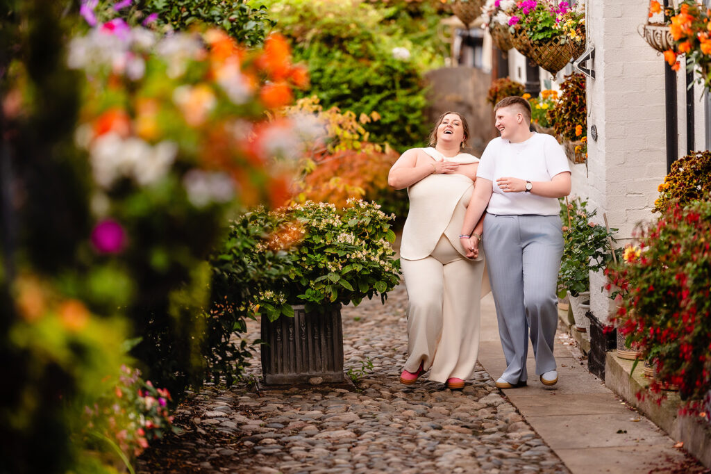 Marianne and Emma walking hand in hand down a cobbled lane lined with flowers in Knutsford