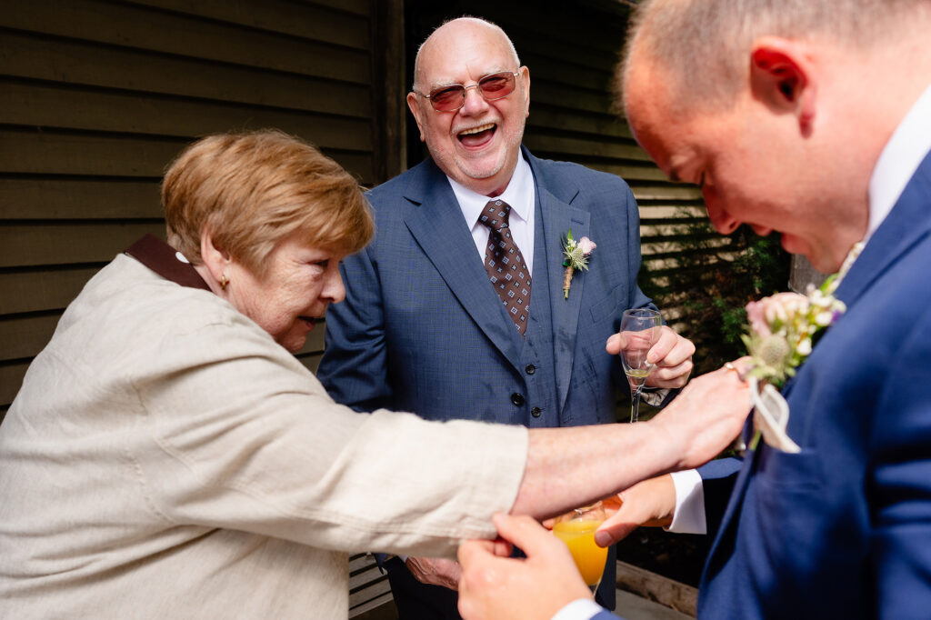 Older guest laughing as a woman adjusts a buttonhole during the drinks reception