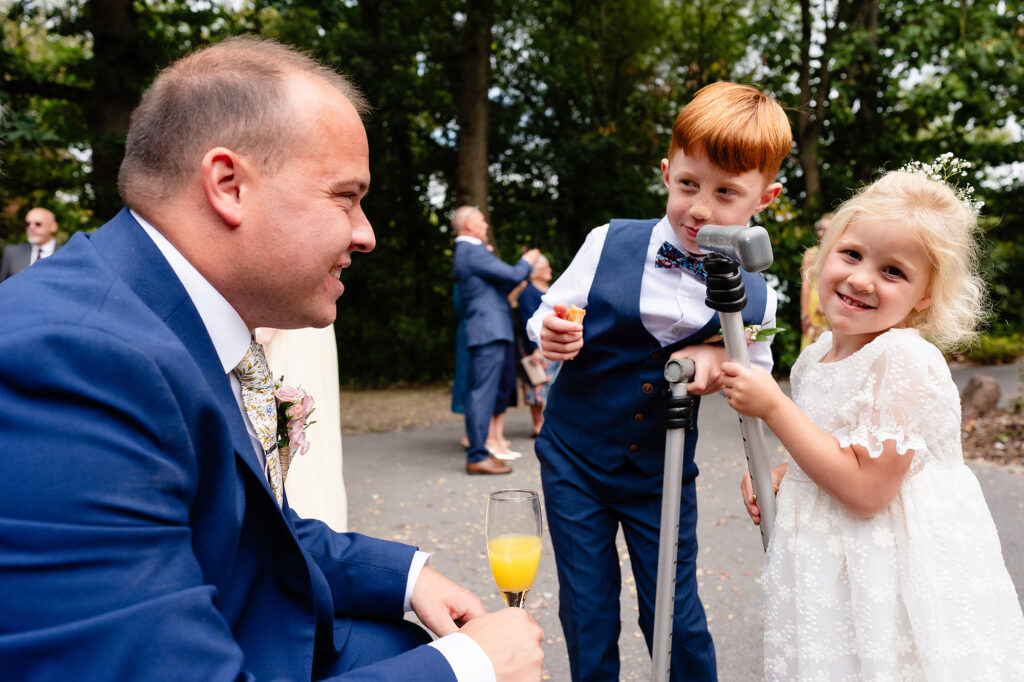 Page boy and flower girl grinning at a guest in a blue suit during the drinks reception