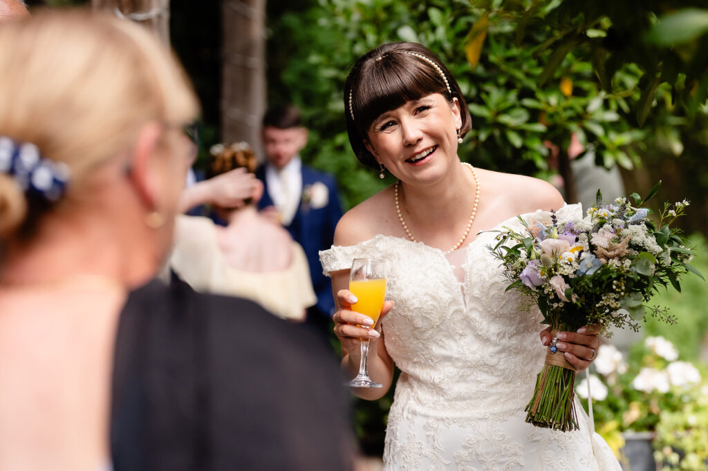Bride laughing with a drink in one hand and her wildflower bouquet in the other in the gardens at The Oak Tree of Peover
