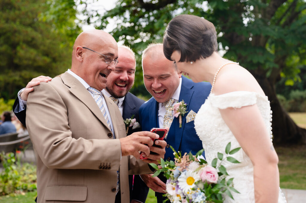 Bride and groom laughing at something on a guest's phone between the group photos