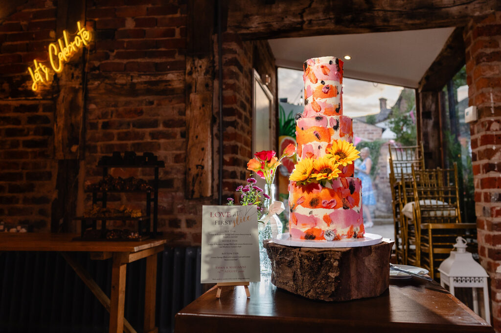 Tiered wedding cake painted in pink, orange and red and topped with sunflowers, by Merryfall Cakes