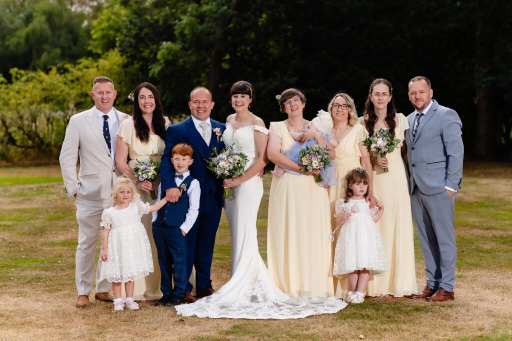 Group with bridesmaids, a page boy and flower girls gathered around the newlyweds on the lawn