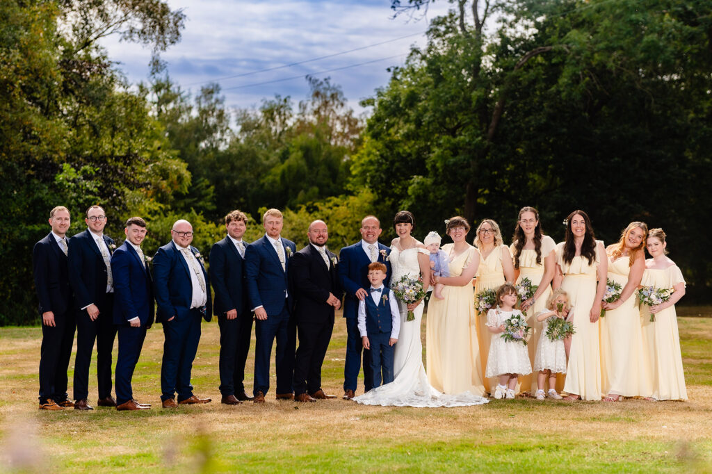 Full bridal party of groomsmen in navy suits and bridesmaids in pale yellow on the lawn at The Oak Tree of Peover