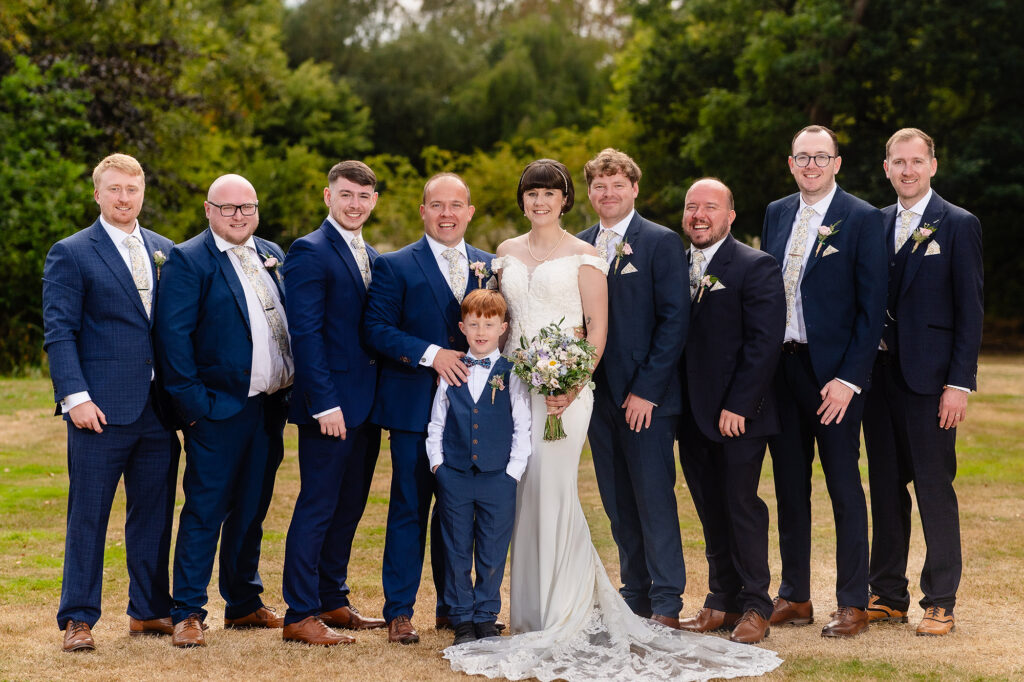 Bride standing with the groom, groomsmen and page boy in navy suits on the lawn