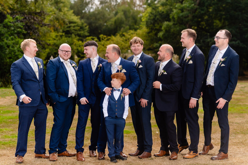 Groom, groomsmen and page boy laughing at each other in a line on the lawn