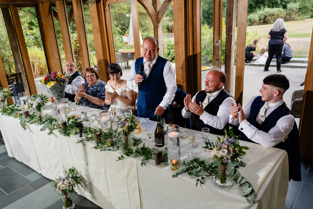 Groom giving his speech at the top table, decorated with eucalyptus and wildflowers, as the table claps