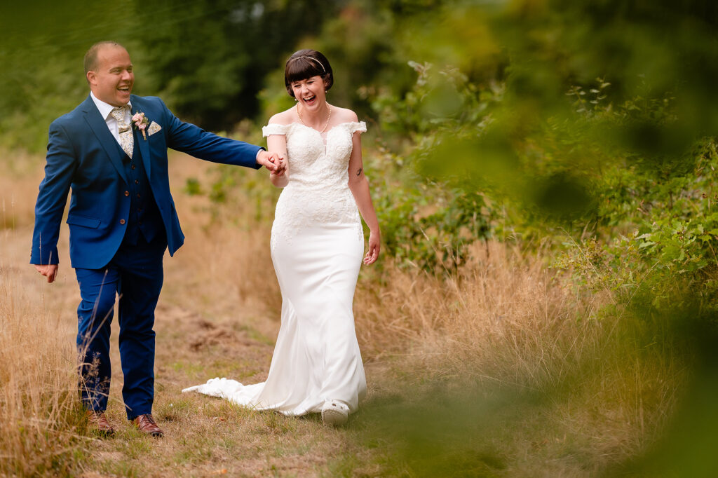 Groom leading the laughing bride by the hand through long golden grass in the grounds of The Oak Tree of Peover